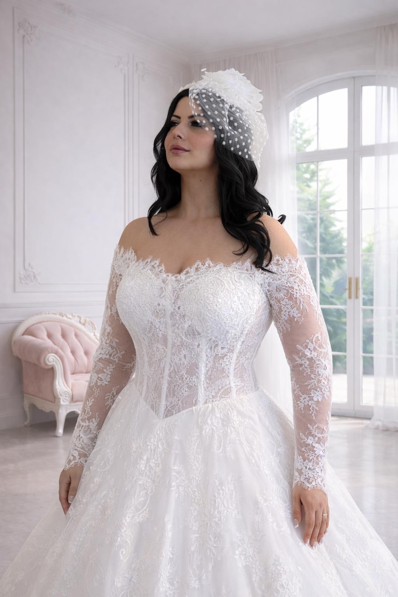 Peut inclure: Une robe de mari&eacute;e blanche &agrave; &eacute;paules d&eacute;nud&eacute;es avec des manches longues en dentelle, un corsage style corset aux d&eacute;tails floraux complexes et une jupe ample. Un bibi blanc assorti avec un voile &agrave; pois est port&eacute; sur la t&ecirc;te.