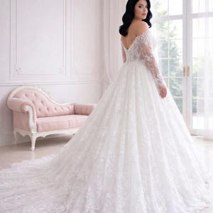 Peut inclure: Une robe de mari&eacute;e blanche &agrave; &eacute;paules d&eacute;nud&eacute;es avec des manches longues en dentelle et une jupe ample arrivant au sol, dot&eacute;e d'une longue tra&icirc;ne. Le corsage et les manches sont orn&eacute;s de broderies florales en dentelle complexe.