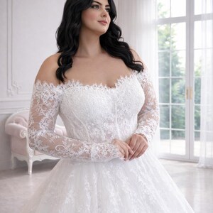 Peut inclure: Une robe de mari&eacute;e blanche &agrave; &eacute;paules d&eacute;nud&eacute;es avec des manches longues en dentelle transparente et un d&eacute;collet&eacute; en c&oelig;ur. Le corsage et la jupe ample sont orn&eacute;s de broderies florales complexes, cr&eacute;ant un aspect textur&eacute; et superpos&eacute; sur la robe.