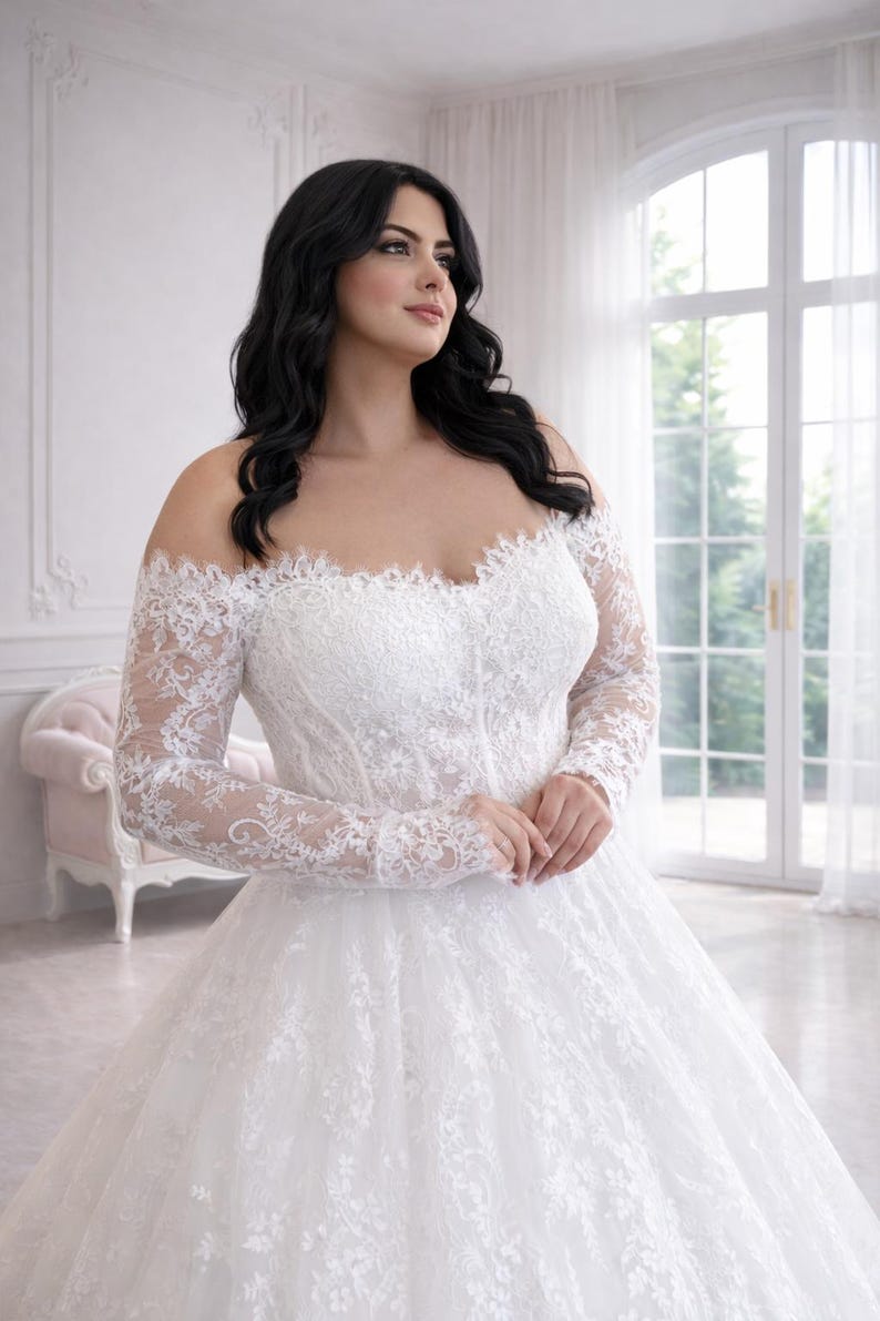 Peut inclure: Une robe de mari&eacute;e blanche &agrave; &eacute;paules d&eacute;nud&eacute;es avec des manches longues en dentelle transparente et un d&eacute;collet&eacute; en c&oelig;ur. Le corsage et la jupe ample sont orn&eacute;s de broderies florales complexes, cr&eacute;ant un aspect textur&eacute; et superpos&eacute; sur la robe.
