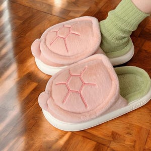 May include: A pair of pink fuzzy slippers shaped like a turtle shell. The slippers have a green lining and a white sole.