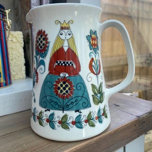 Può includere: Una brocca in ceramica bianca con una colorata illustrazione di una figura coronata dai lunghi capelli biondi, che indossa un top rosso e una gonna verde acqua. Il design include girasoli stilizzati, motivi floreali e un bordo decorativo.