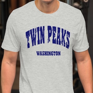May include: A light grey t-shirt with the text "TWIN PEAKS WASHINGTON" in dark blue. The shirt is short-sleeved and appears to be made of a soft material. The person is standing in a gym.