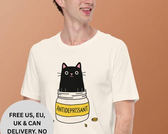 Könnte beinhalten: Cremefarbenes T-Shirt mit einer Cartoon-Katze in einem gelb-weißen Glas mit der Aufschrift "ANTIDEPRESSANT". Das Design enthält gelbe Pillen-Illustrationen. Das T-Shirt wird von einer Person getragen.
