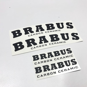 Op de afbeelding: Twee witte rechthoekige stickers met het woord "BRABUS" in vette zwarte letters, met "CARBON CERAMIC" eronder. De ene sticker is groter dan de andere. De stickers liggen op een witte ondergrond.