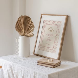 K&ouml;nnte beinhalten: Ein gerahmtes Schild mit dem Text "CARDS and GIFTS" und einer Cowboyhut-Illustration. Das Schild befindet sich in einem Holzrahmen, der an einer wei&szlig;en Wand lehnt. Eine wei&szlig;e Vase mit einem getrockneten Palmblatt und zwei B&uuml;cher mit einem goldenen Armband liegen auf einer wei&szlig;en Tischdecke.