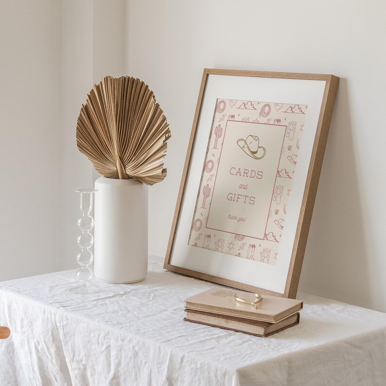 K&ouml;nnte beinhalten: Ein gerahmtes Schild mit dem Text "CARDS and GIFTS" und einer Cowboyhut-Illustration. Das Schild befindet sich in einem Holzrahmen, der an einer wei&szlig;en Wand lehnt. Eine wei&szlig;e Vase mit einem getrockneten Palmblatt und zwei B&uuml;cher mit einem goldenen Armband liegen auf einer wei&szlig;en Tischdecke.