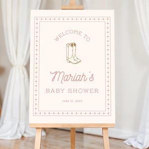 K&ouml;nnte beinhalten: Ein cremefarbenes Schild auf einer Holzstaffelei mit dem Text "Welcome to Mariah's Baby Shower" in einer Mischung aus Schriftarten. Das Schild enth&auml;lt eine Illustration von Cowboy-Stiefeln und das Datum 21. Juni 2025.