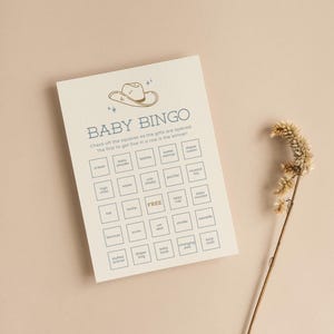May include: A beige baby shower bingo card with a blue cowboy hat illustration at the top. The card has a grid of 25 squares with baby-related items listed in each square, such as onesie, baby powder, wipes, bottle warmer, diaper cream, high chair, crib sheets, pacifier, musical toy, hat, bottle, free, sippy cup, baby monitor, bouncer, socks, car seat, stroller, swaddle, stuffed animal, diaper bag, baby book, changing pad, and burp cloth.