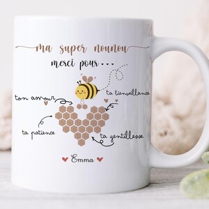 May include: White ceramic mug with a bee and honeycomb design. The mug has the text "ma super nounou merci pour..." and other French text. The mug is on a white surface with a woven basket and greenery in the background.