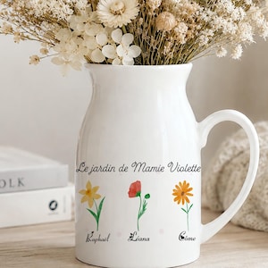 Puede incluir: Una jarra de cerámica blanca con el texto Le jardin de Mamie Violette y tres pequeñas ilustraciones florales con los nombres Raphaël, Liana y Côme debajo. La jarra está llena de un gran arreglo de flores secas de color beige.