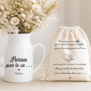 Peut inclure: Un pichet en céramique blanche avec anse, portant l'inscription noire "Parrain pour la vie..." et un petit cœur rouge, rempli de fleurs séchées beiges et blanches, accompagné d'une pochette à cordon beige avec du texte noir et un cœur.