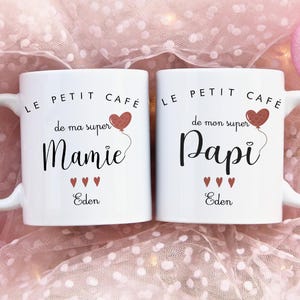 Peut inclure: Deux mugs en céramique blanche avec l'inscription noire "LE PETIT CAFÉ de ma super Mamie" et "LE PETIT CAFÉ de mon super Papi". Chaque mug a un motif de ballon cœur rouge et le nom "Eden". Décorations de cœur roses en arrière-plan.