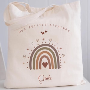 Puede incluir: Bolso tote color crema con el texto "MES PETITES AFFAIRES" sobre un diseño de arcoíris en tonos marrón, verde y naranja. El nombre "Jade" está escrito debajo. El bolso tiene dos asas.