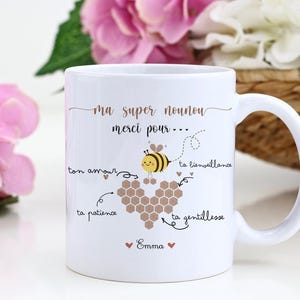 May include: White ceramic mug with a handle, featuring a bee illustration and the text "ma super nounou merci pour..." in gold lettering. The design includes a heart-shaped honeycomb and the name "Emma".