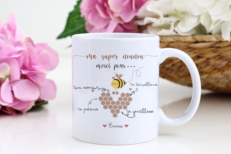May include: White ceramic mug with a handle, featuring a bee illustration and the text "ma super nounou merci pour..." in gold lettering. The design includes a heart-shaped honeycomb and the name "Emma".