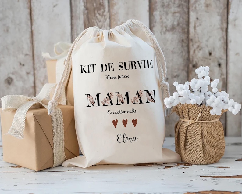 May include: A cream-colored drawstring bag with the text "KIT DE SURVIE D'une future MAMAN Exceptionnelle" in black and floral lettering. Below the text are three brown hearts and the name "Elora". The bag is next to wrapped gifts and a burlap vase with white flowers.