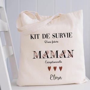 May include: A cream-colored tote bag with black text that reads "KIT DE SURVIE" and "MAMAN Exceptionnelle Elora". The word "MAMAN" has floral accents. Three brown hearts are below the word "Exceptionnelle".