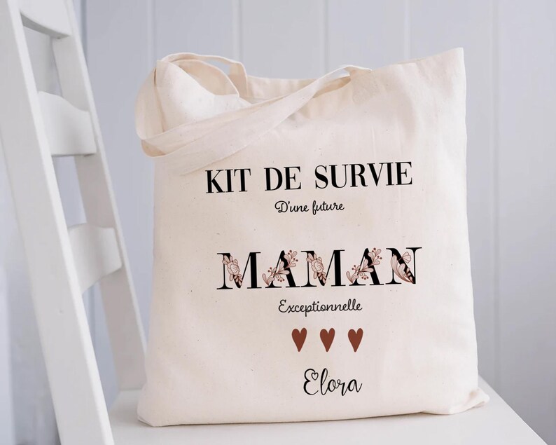 May include: A cream-colored tote bag with black text that reads "KIT DE SURVIE" and "MAMAN Exceptionnelle Elora". The word "MAMAN" has floral accents. Three brown hearts are below the word "Exceptionnelle".