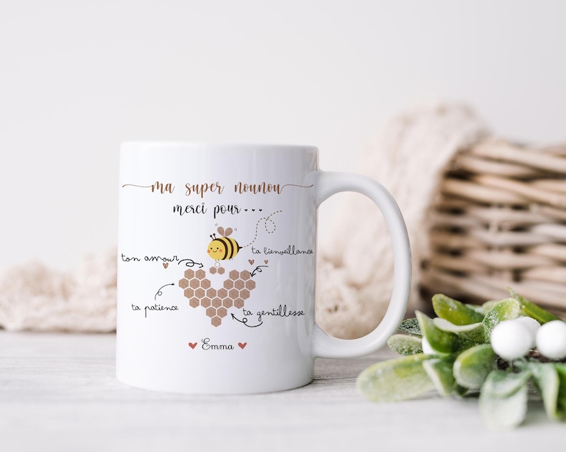 May include: White ceramic mug with a bee and honeycomb design. The mug has the text "ma super nounou merci pour..." and other French text. The mug is on a white surface with a woven basket and greenery in the background.