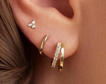 May include: Close-up of a person's ear adorned with multiple gold earrings. A small stud earring with three clear stones is at the top. Below, a small gold hoop and a double hoop earring with clear stones are visible.