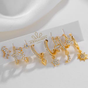May include: A collection of gold-coloured earrings featuring star, moon, and geometric designs, embellished with clear gemstones. The earrings are presented on a white card, with the text "JEWELLOSITY LONDON" visible. The set includes various hoop and stud styles.