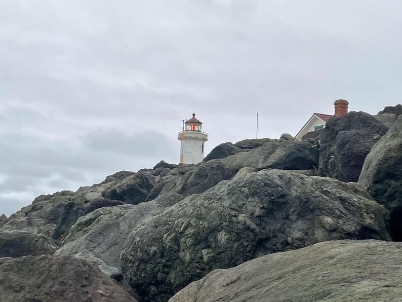May include: A white lighthouse with an orange roof stands on a rocky cliff overlooking the ocean. The sky is overcast and gray.