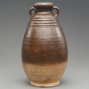 Könnte beinhalten: Eine braune Keramikvase mit rundem Körper und zwei kleinen Griffen in der Nähe der Oberseite. Die Vase hat ein zweifarbiges Farbschema, mit einem dunkleren braunen oberen Teil und einem helleren braunen unteren Teil. Die Vase hat eine strukturierte Oberfläche.