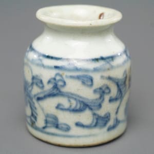 May include: Small, white ceramic jar with blue floral patterns. The jar has a rounded body and a narrow opening. The blue design wraps around the jar's midsection, creating a decorative band. The jar is likely a decorative item or a small container.
