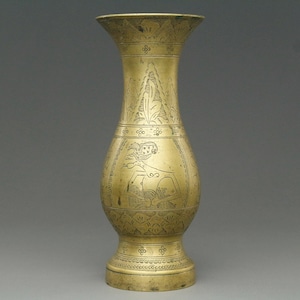 Op de afbeelding: Een hoge, antieke messing vaas met ingewikkelde geëtste ontwerpen. De vaas heeft een uitlopende bovenkant, een bolvormig middengedeelte met een gedetailleerde figuur en een voetstuk. Het oppervlak is versierd met bloemen en geometrische patronen. Ongeveer 25 cm hoog.