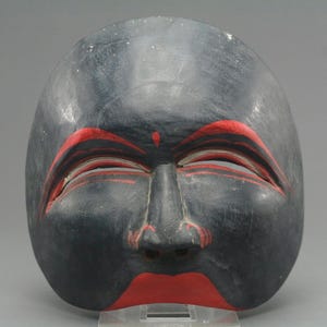 May include: A black wooden mask with red detailing. The mask has red eyebrows, a small red mark on the forehead, and red lips. The eyes are partially closed, and the mask has a calm expression. The mask is likely used for theatrical or decorative purposes.
