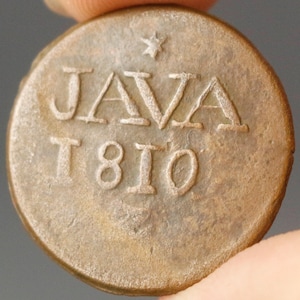 Puede incluir: Una moneda circular y desgastada, de color bronce, con la palabra "JAVA" y el año "1810". Una pequeña estrella está encima de "JAVA". La moneda muestra signos de desgaste y el texto está en relieve.