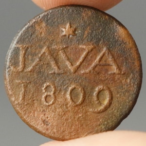 Op de afbeelding: Een ronde, antieke bronzen munt met het woord "JAVA" en het jaartal "1809" erin geëmbosseerd. Boven het woord "JAVA" staat een kleine ster. De munt heeft een verweerde uitstraling.