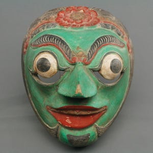 Puede incluir: Una máscara de madera tallada a mano con una cara verde, labios rojos y ojos grandes y expresivos. La máscara presenta un diseño floral rojo en la frente y probablemente se utiliza con fines ceremoniales o decorativos.