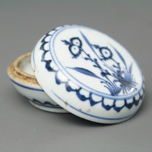 Puede incluir: Pequeña caja de baratijas redonda de cerámica blanca con tapa. La tapa está decorada con un diseño floral azul y un borde festoneado. El interior de la caja es visible, mostrando un borde marrón claro.