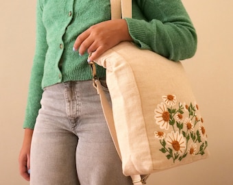 May include: A beige canvas tote bag with a long strap and an embroidered design of white daisies with brown centers and green leaves.