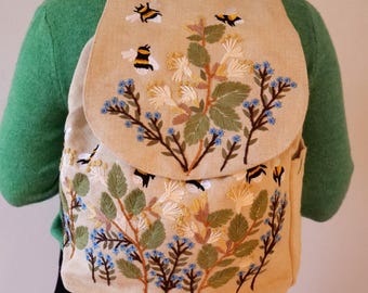 May include: Beige backpack with embroidered floral and bee design. The embroidery features green leaves, blue flowers, and yellow and black bees. The backpack has a top handle and adjustable straps.