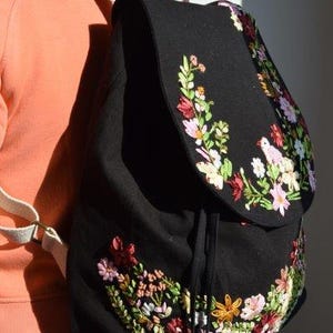 Pode incluir: Uma mochila de lona preta com bordado floral em tons de rosa, vermelho e verde. A mochila tem fecho de cord&atilde;o e al&ccedil;as ajust&aacute;veis.