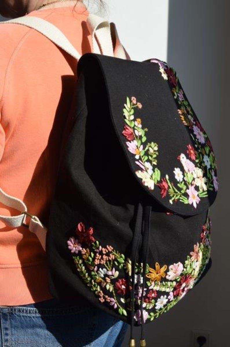 Pode incluir: Uma mochila de lona preta com bordado floral em tons de rosa, vermelho e verde. A mochila tem fecho de cord&atilde;o e al&ccedil;as ajust&aacute;veis.