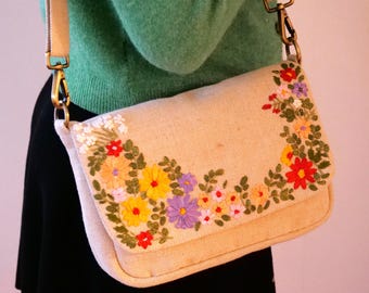 May include: A beige canvas crossbody bag with a floral embroidery design. The embroidery features a variety of colorful flowers, including yellow, red, purple, and white.