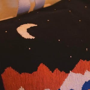 May include: Black fabric embroidered with a night sky scene. A crescent moon and scattered stars are stitched in white. The bottom features a mountain range in orange, white, and blue embroidery. The item appears to be a pillow or tote bag.