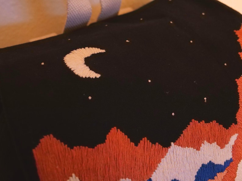 May include: Black fabric embroidered with a night sky scene. A crescent moon and scattered stars are stitched in white. The bottom features a mountain range in orange, white, and blue embroidery. The item appears to be a pillow or tote bag.