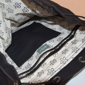 Pode incluir: Mochila de lona preta com fecho de cord&atilde;o e um bolso com z&iacute;per. O forro interior &eacute; branco com um padr&atilde;o floral e o texto "The Baguette" &eacute; vis&iacute;vel no bolso com z&iacute;per.