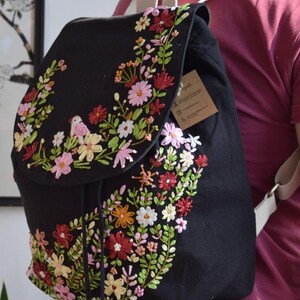 Pode incluir: Mochila de lona preta com um design de bordado floral. O bordado apresenta flores rosa, vermelhas, amarelas e verdes, bem como um pequeno p&aacute;ssaro.