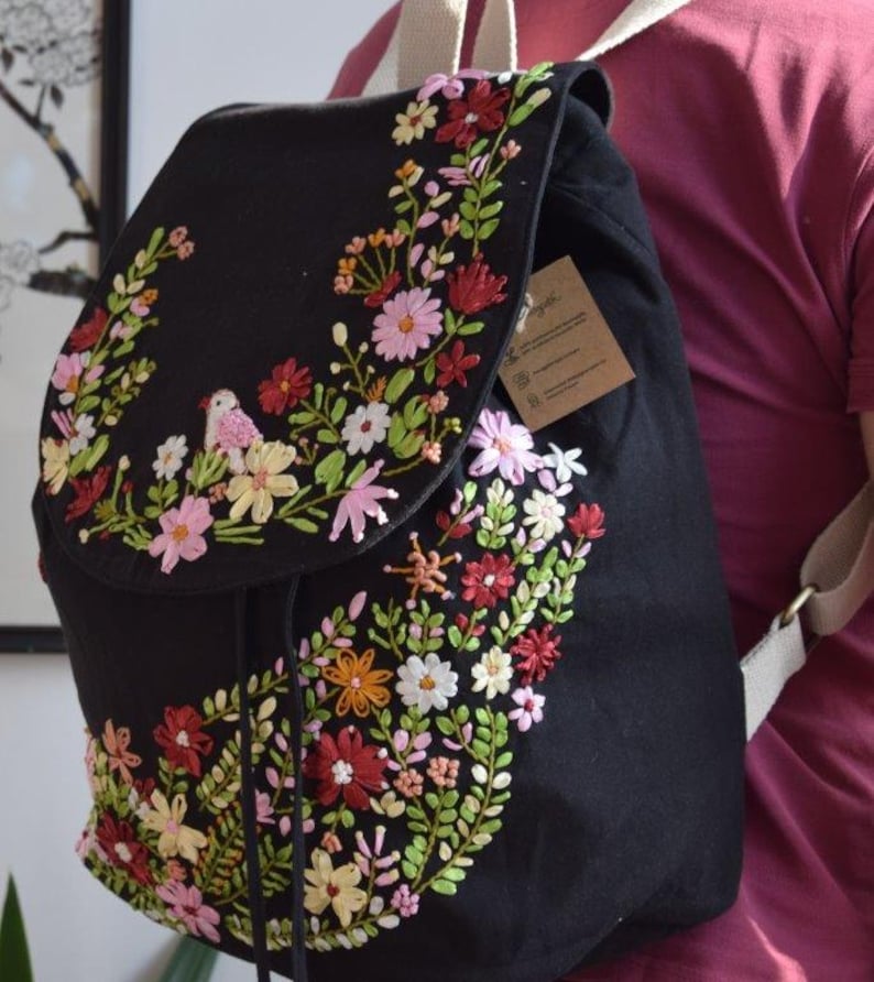 Pode incluir: Mochila de lona preta com um design de bordado floral. O bordado apresenta flores rosa, vermelhas, amarelas e verdes, bem como um pequeno p&aacute;ssaro.