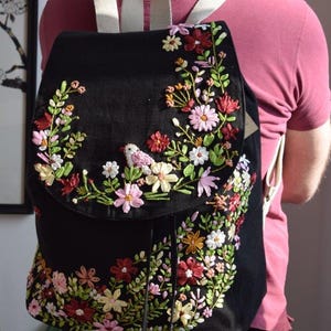 May include: A black canvas backpack with a floral embroidery design. The embroidery features a variety of colorful flowers, leaves, and a small bird.
