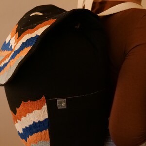 May include: Black backpack with embroidered mountain range design in orange, white, blue, and gray. The backpack has a flap closure and a small front pocket. White shoulder straps are visible.