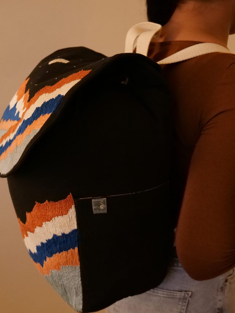 May include: Black backpack with embroidered mountain range design in orange, white, blue, and gray. The backpack has a flap closure and a small front pocket. White shoulder straps are visible.