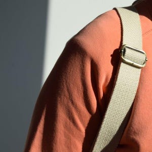 Pode incluir: Um close-up de uma pessoa vestindo uma camisa cor coral com uma al&ccedil;a de lona bege. A al&ccedil;a tem uma fivela de lat&atilde;o.