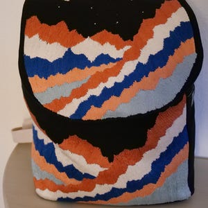 May include: Black backpack with embroidered mountain range design in orange, blue, white, and gray. The top flap features a crescent moon and stars. The backpack has a beige handle and straps.